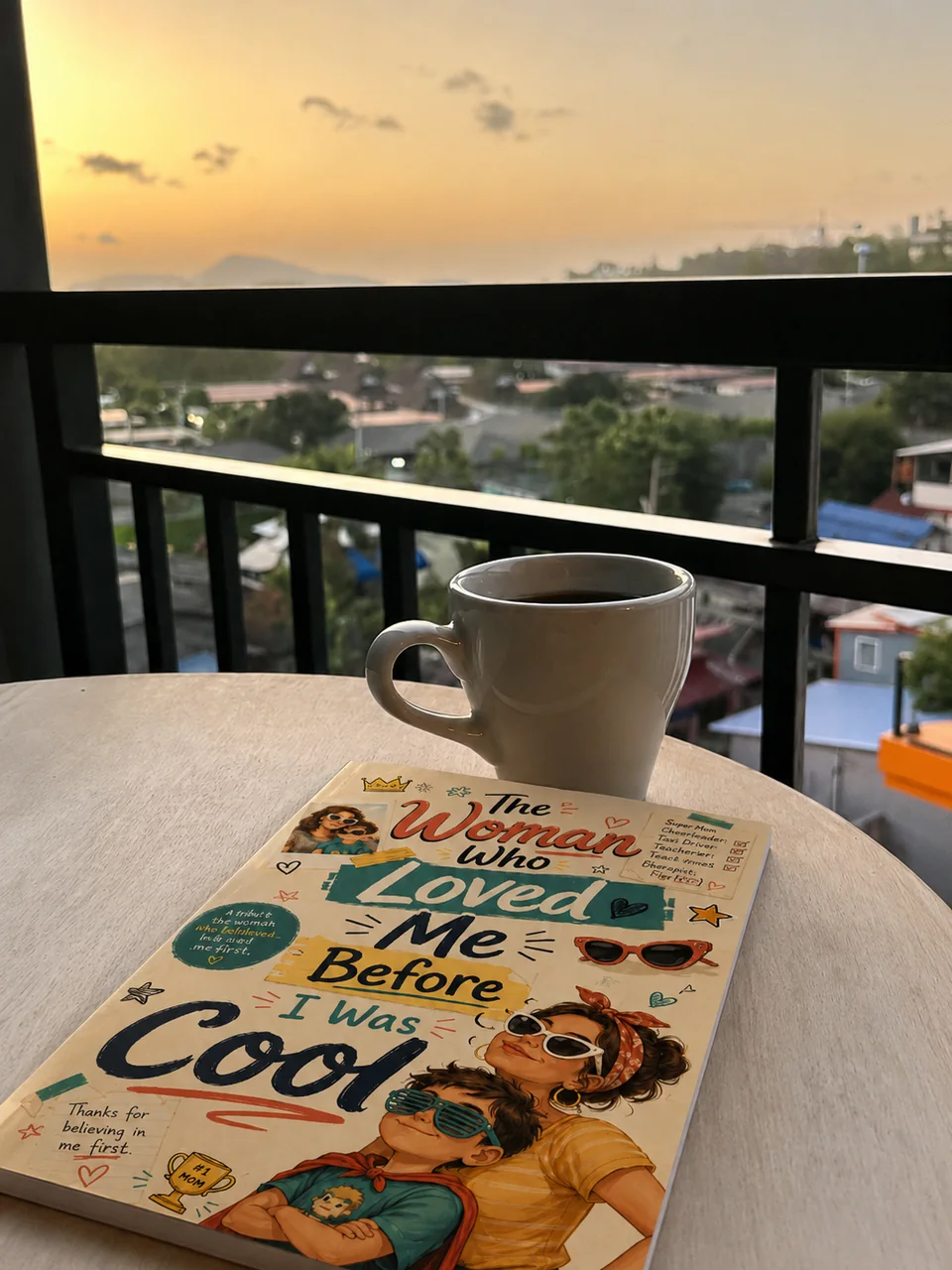 A Keepsake softcover titled The Woman Who Loved Me Before I Was Cool, on a balcony table beside a cup of coffee at sunset